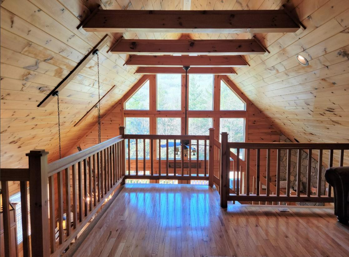The loft area features high vaulted ceilings made of natural wood, with large windows that provide a bright view of the surrounding trees. Wooden railings outline the open space, enhancing the cabin's rustic charm and providing a sense of openness.