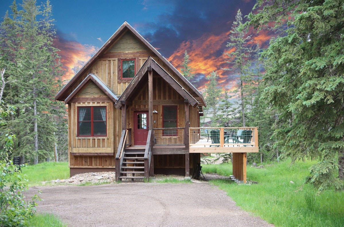 The cabin features a rustic exterior with wooden siding and a sloped roof. A spacious deck is located at the front, surrounded by lush greenery. Large windows allow for natural light, and the welcoming entryway is accentuated by steps leading to the door.