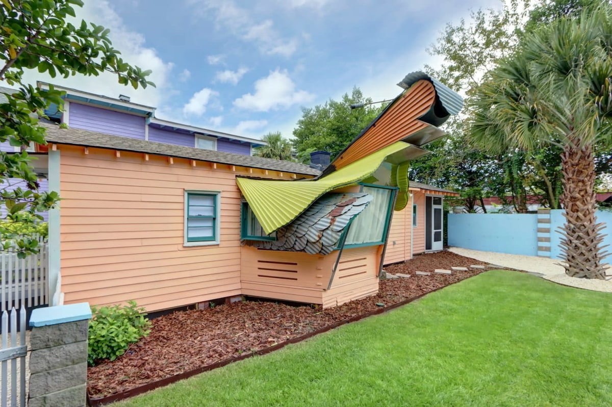 The exterior of a vibrant, uniquely designed house is showcased, featuring an unconventional roof that appears to playfully extend outward. Surrounding greenery includes well-maintained grass and palm trees, while the colorful finish adds a distinct character to the property.