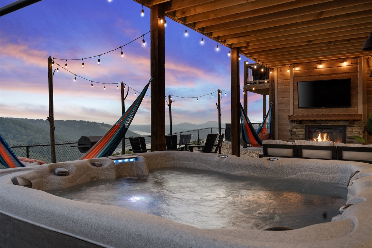 An inviting hot tub is situated under a wooden pergola, surrounded by vibrant hammocks and a cozy seating area. Beautiful outdoor lights illuminate the space, casting a warm glow as the serene hills are visible in the distance, creating a perfect backdrop for relaxation.