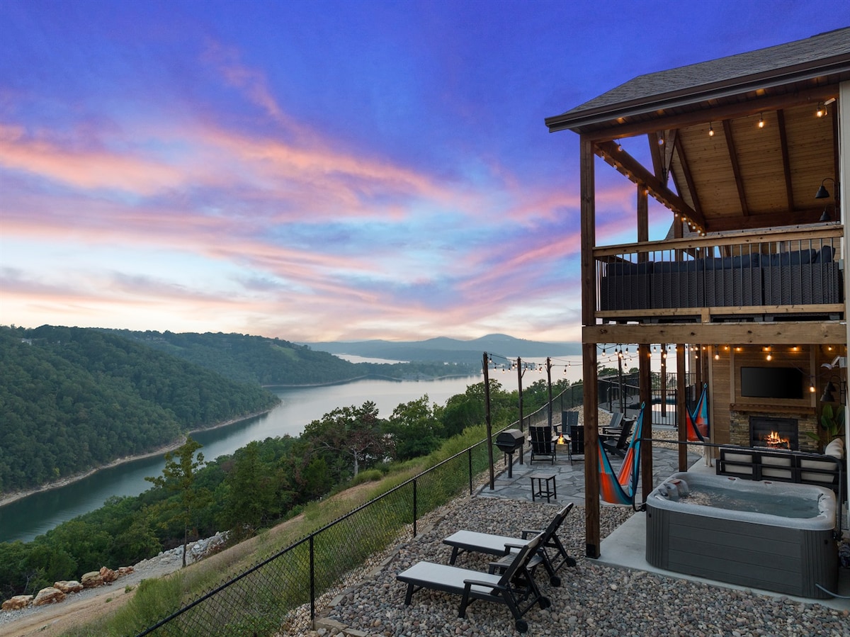 A scenic view of Table Rock Lake is presented from an expansive outdoor deck. The image includes a hot tub, a seating area with a firepit, and colorful hammocks. The cabin features warm lighting along the deck, enhancing the serene landscape.
