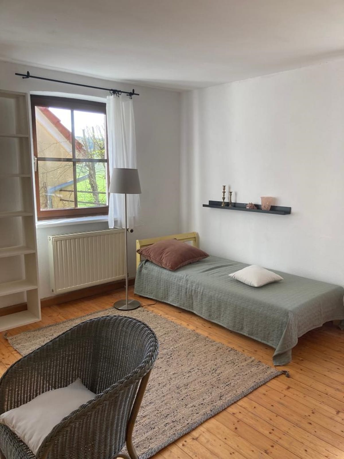 A simple bedroom features a single bed covered with a muted green blanket and a small white pillow. A wooden shelf displays decorative items. A comfortable chair is positioned near a window, which is dressed with sheer curtains, allowing natural light to fill the space.