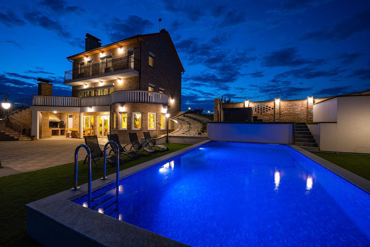 The villa is showcased at dusk, illuminated by outdoor lights. A shimmering blue pool is visible in the foreground, with steps leading down from the patio. The three-story structure features several spacious balconies, enhancing the tranquil setting surrounded by nature.