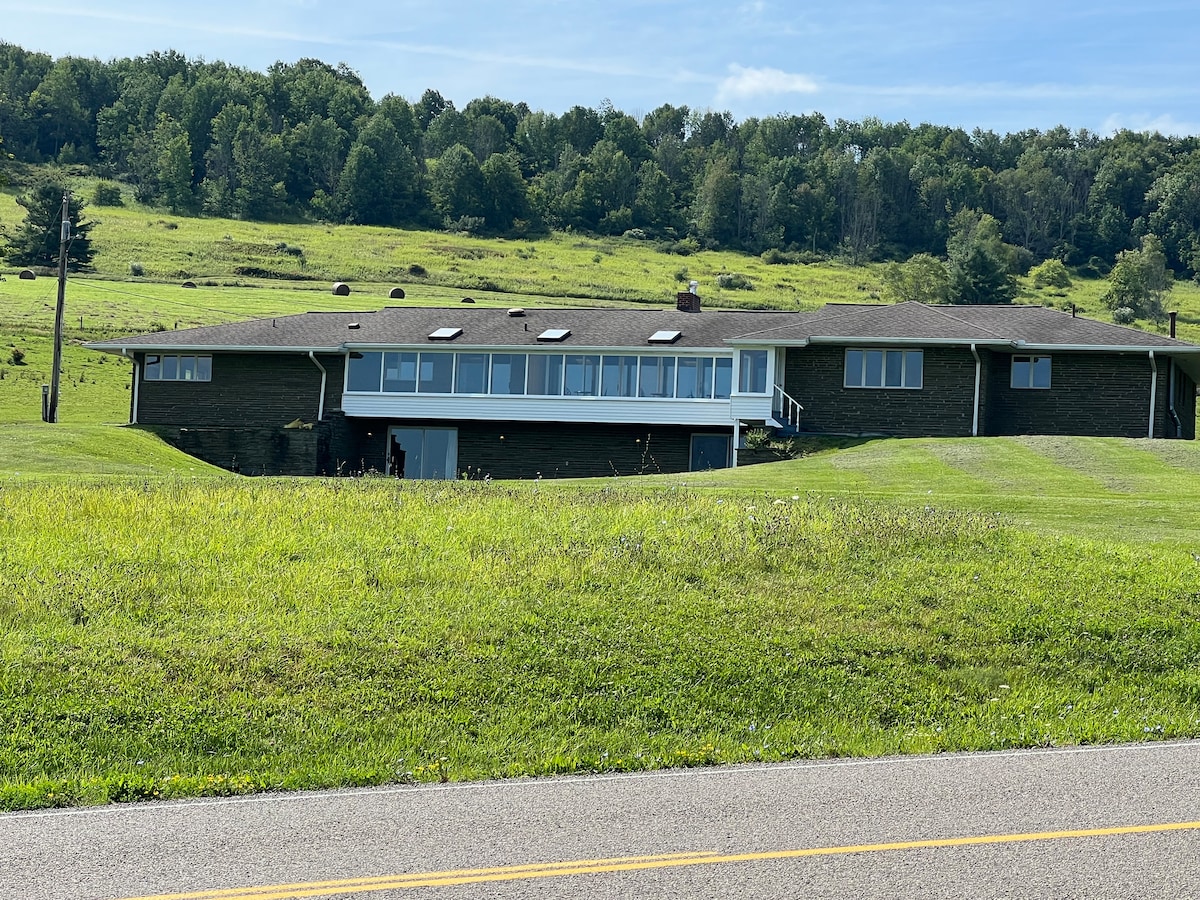 The exterior of the Sun Room Apartment is displayed, featuring a sprawling, single-story structure with a long row of wide windows. Lush green grass surrounds the building, with gentle hills and trees visible in the background under a clear blue sky.