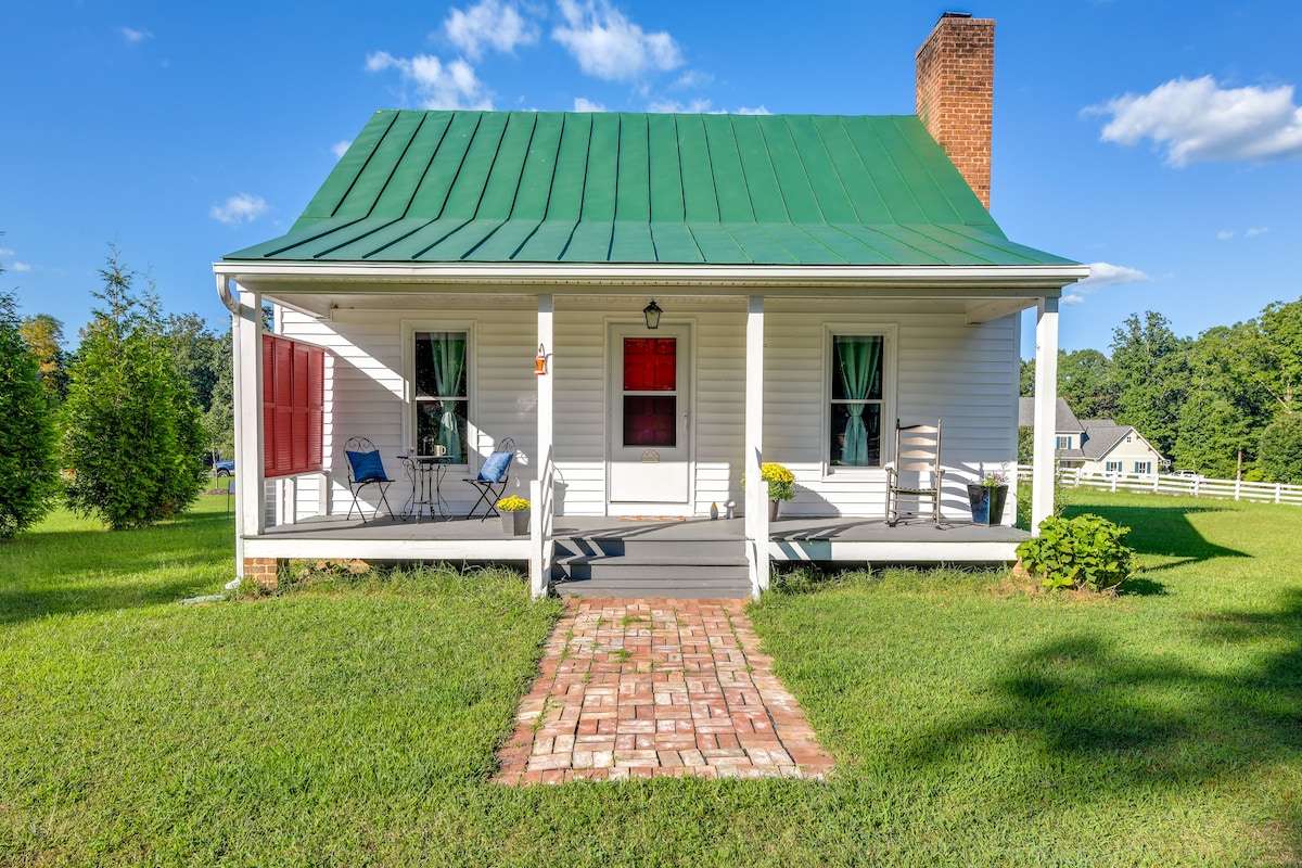 A charming cottage exterior is depicted, featuring a green metal roof and a welcoming front porch. Two blue chairs and a wooden rocking chair are arranged on the porch, with a brick pathway leading to the door. Lush green grass surrounds the property.
