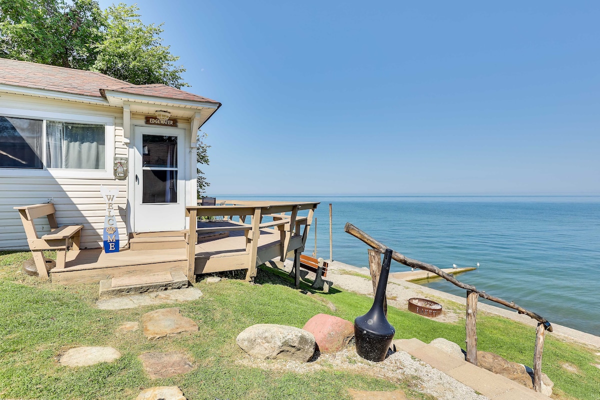 The entrance of the cottage is shown, featuring a wooden deck that leads to a welcoming door. A handcrafted sign is visible, and a bench is positioned nearby. The serene lake extends towards the horizon, complemented by a clear blue sky.