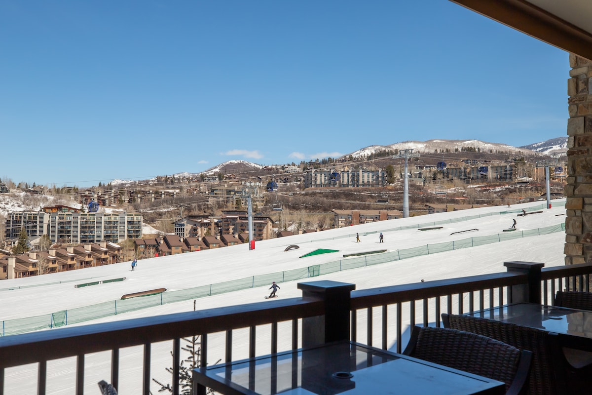 A winter landscape is captured from a balcony view, with ski slopes visible in the foreground. The clear blue sky complements the snowy terrain, where skiers and snowboarders can be seen enjoying the slopes. Nearby structures are partially visible in the background.