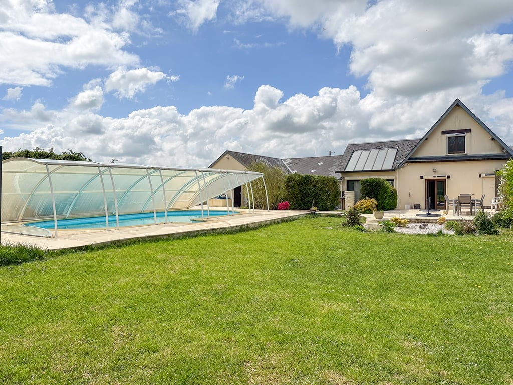 A spacious garden is visible, featuring a heated swimming pool covered by a transparent dome. The house stands nearby, surrounded by greenery, with a patio area that includes outdoor seating. The sky above is partly cloudy, contributing to a bright and inviting atmosphere.