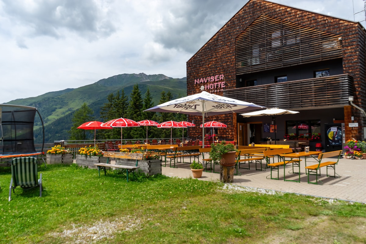 The exterior of the Naviser Hütte showcases a rustic building with wooden cladding. Colorful parasols shade spacious outdoor seating, surrounded by vibrant flower beds. A view of the surrounding mountains and greenery enhances the tranquil atmosphere of the retreat.