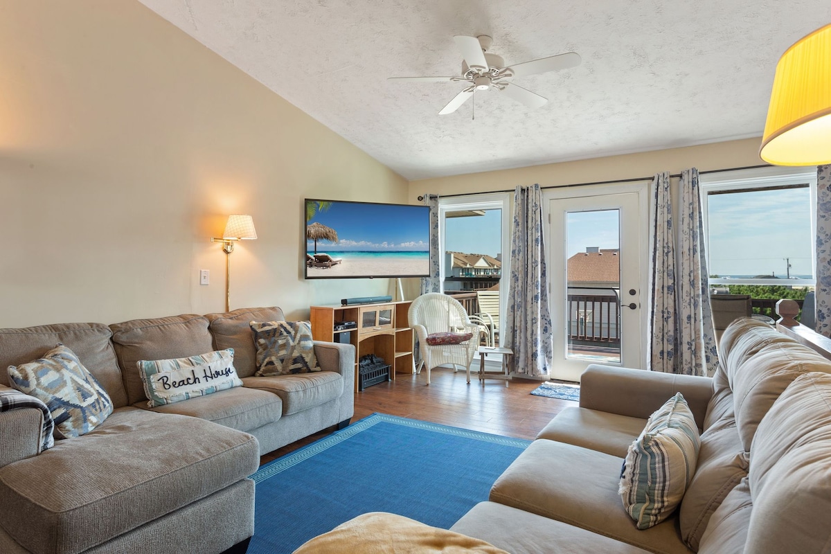 A spacious living area features comfortable seating arrangements on a sectional sofa and a love seat. A large television is mounted on the wall, and natural light fills the space through multiple windows. A doorway leads to an outdoor balcony with views.