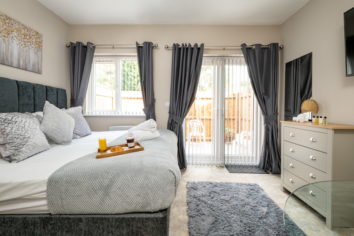 A spacious bedroom features a super-king size bed adorned with soft linens. Natural light streams in through sliding glass doors, leading to a private outdoor area. A comfortable seating area is present, complemented by a chest of drawers and a stylish rug on the floor.