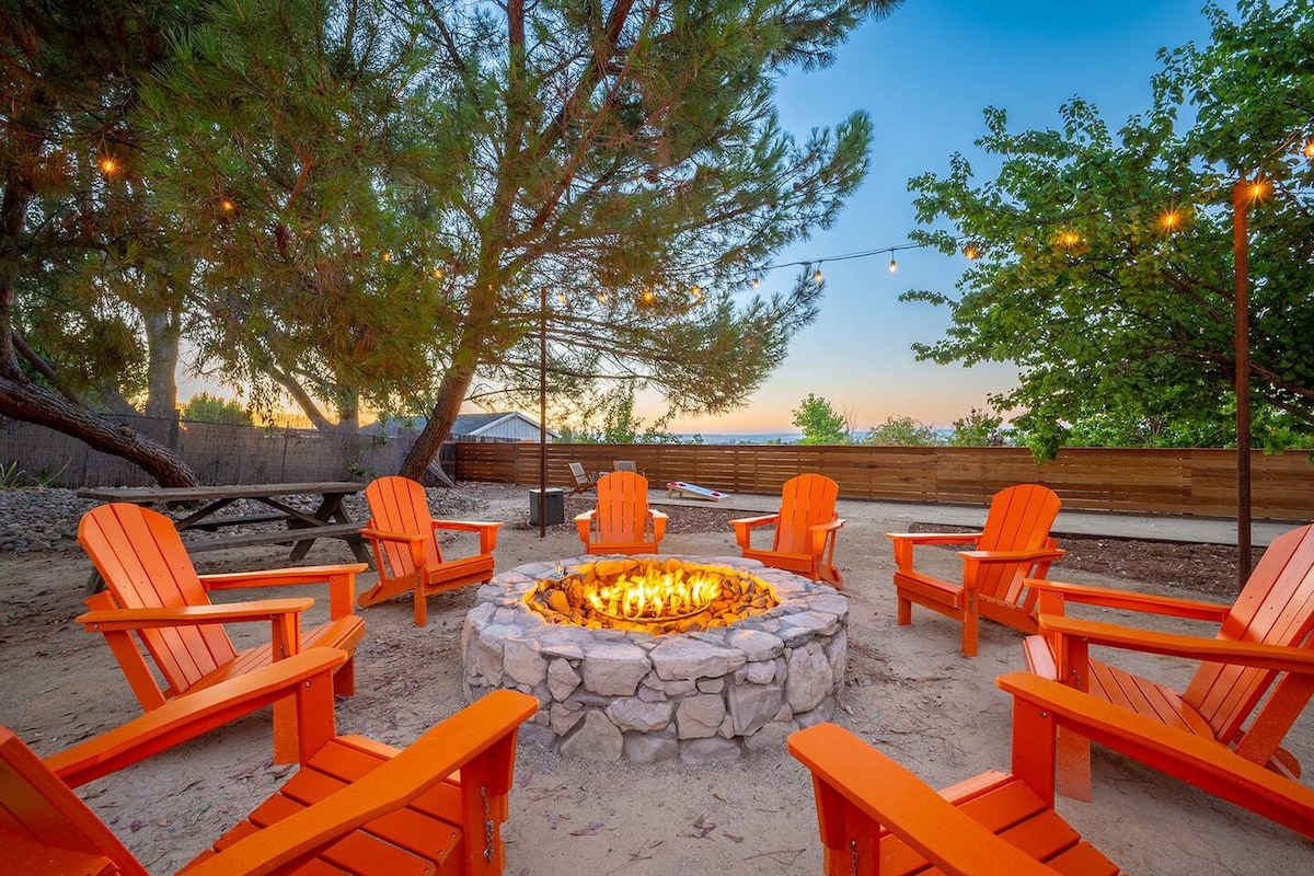 An outdoor fire pit area features eight bright orange Adirondack chairs arranged around a stone fire pit. String lights hang above the seating, illuminating the space softly. Lush greenery and a wooden fence create a private and welcoming setting as dusk approaches.