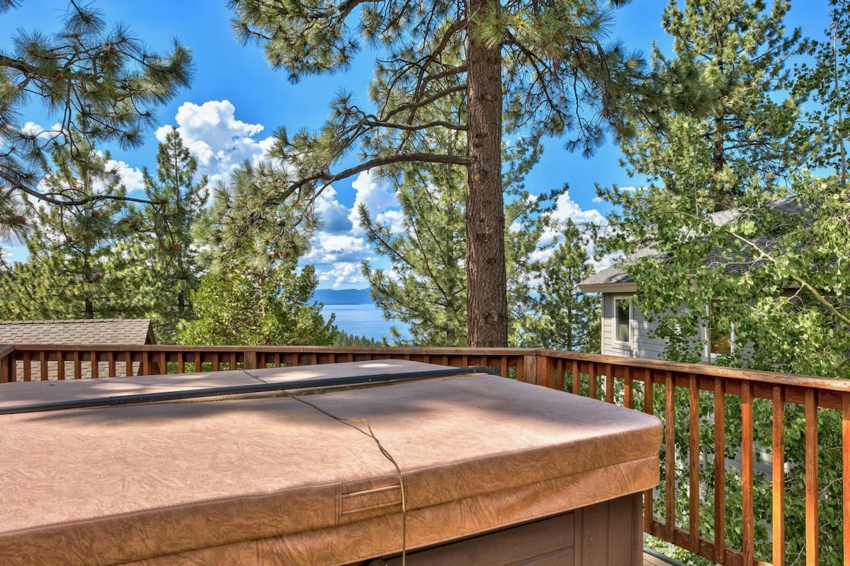 A hot tub with a protective cover is shown on a wooden deck surrounded by tall pine trees. Clear blue skies and fluffy clouds are visible in the background, framing a scenic view of the distant lake.
