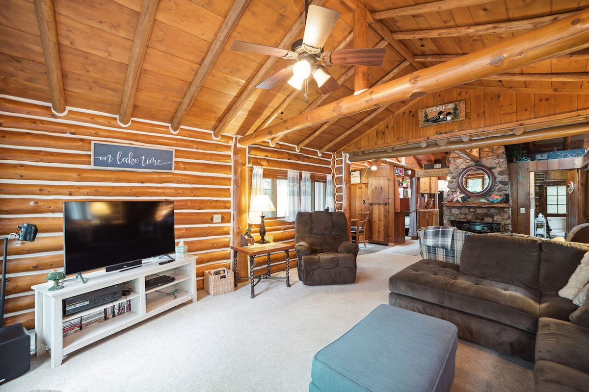 A spacious living area features high wooden ceilings, highlighting the log cabin's rustic charm. Comfortable seating, including a sectional sofa and recliner, invites relaxation, while a wall-mounted television provides entertainment options. Natural light flows through multiple windows, enhancing the cozy ambiance.