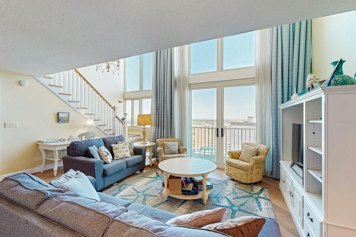 A bright living area highlights a spacious sofa and two armchairs, all arranged around a circular coffee table. Large windows allow abundant natural light to fill the space, while the staircase with an ornate chandelier adds an elegant touch.