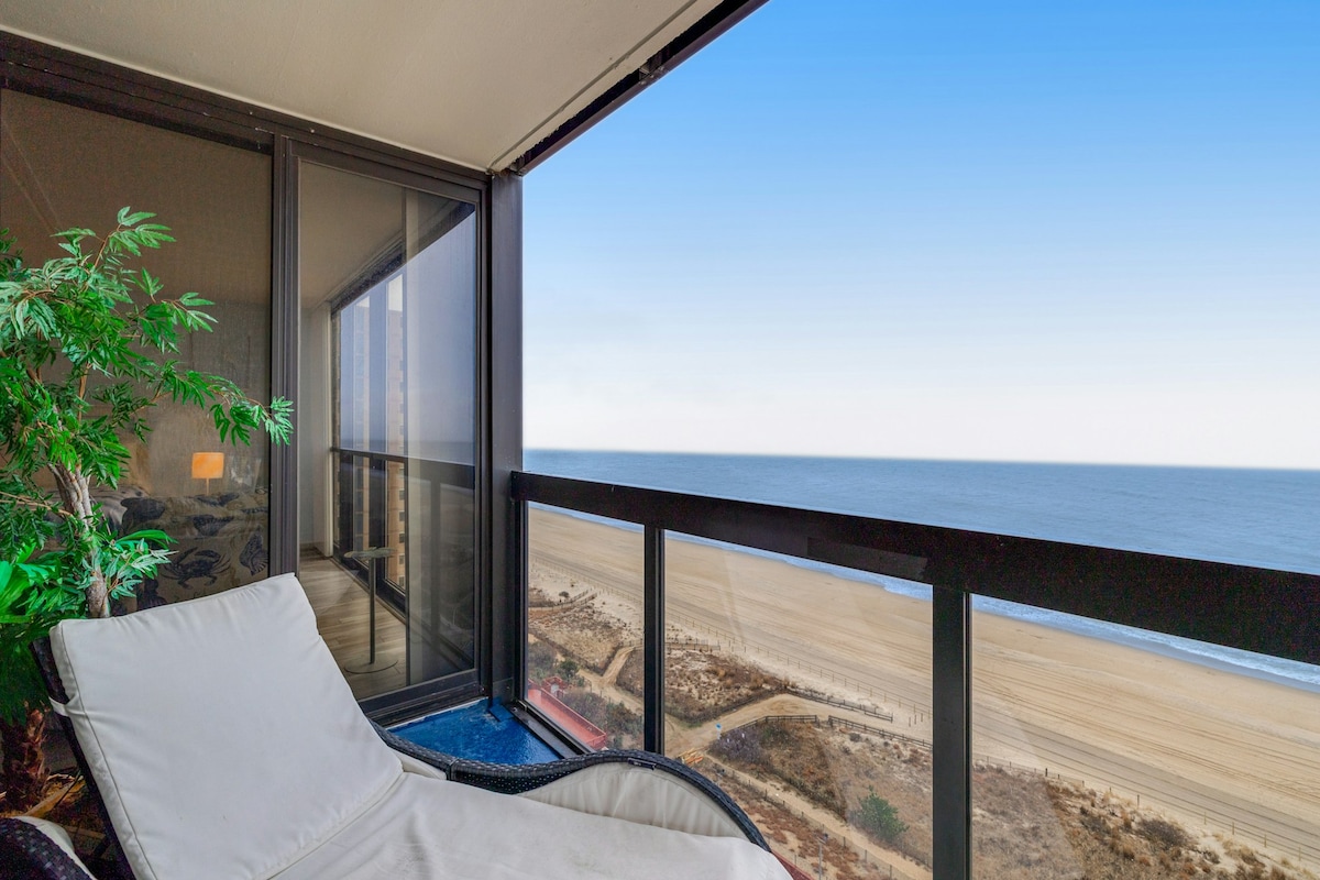 A private balcony offers an unobstructed view of the ocean along with a spacious chaise longue beach chair. The glass railings enhance visibility, while the beach and waves can be seen in the background. Green plant foliage adds a touch of nature to the space.