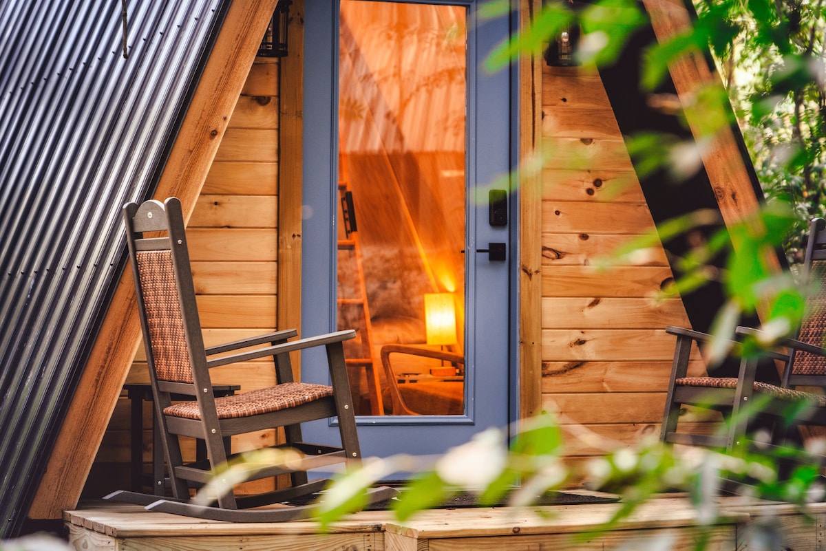 A wooden A-frame cabin entrance is framed by lush green foliage. Rocking chairs are positioned on a spacious front deck, inviting relaxation. Warm light glows softly through the cabin door, suggesting a cozy interior after a day in nature.