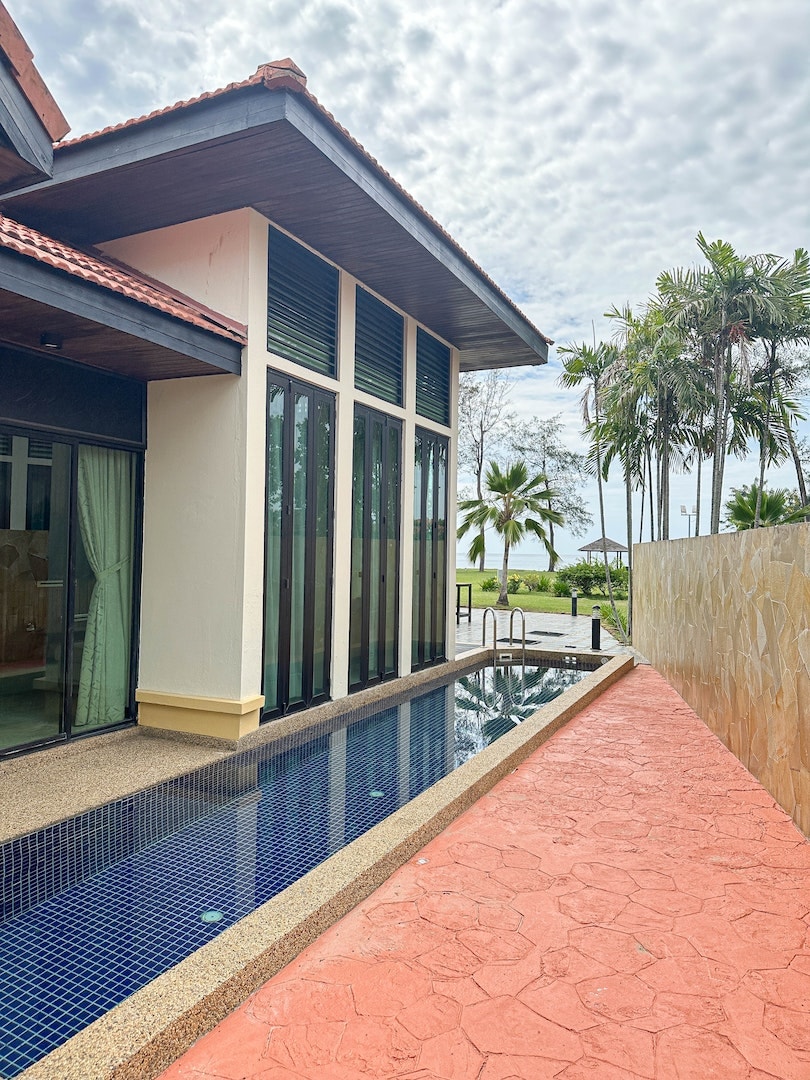 The exterior of the villa features modern architectural elements with large glass panels and a private swimming pool. A stone pathway leads alongside the pool, with lush greenery visible in the background, providing a serene view of the ocean.