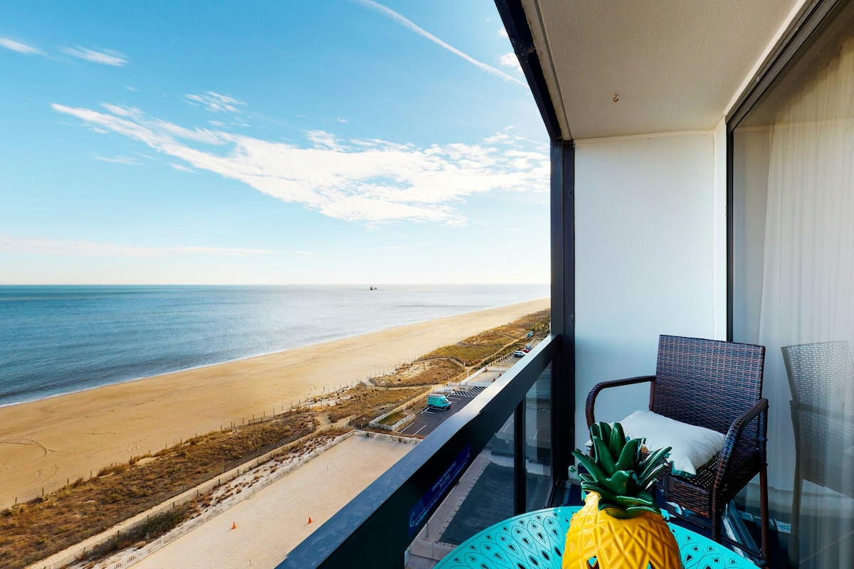 A private balcony overlooks a sandy beach and the Atlantic Ocean with a clear blue sky above. A small table topped with a decorative pineapple and a comfortable chair create a relaxing outdoor space.