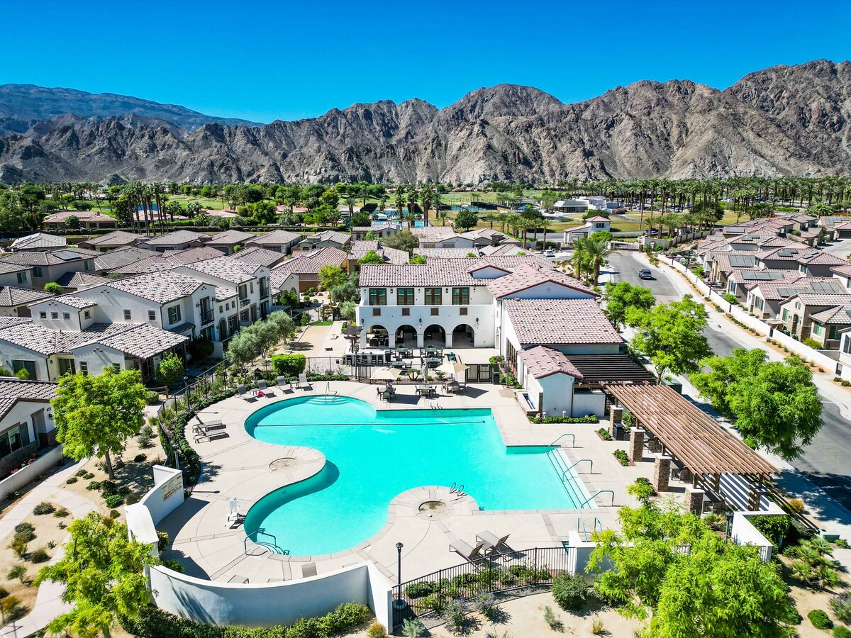 An aerial view captures the expansive community pool with a unique shape, surrounded by lush landscaping and palm trees. Mountain views form a stunning backdrop, highlighting the serene environment of the PGA West Signature community.