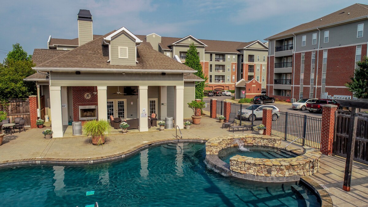 A spacious pool area features a large swimming pool with a waterfall feature and a cozy seating area under a covered patio. Surrounding greenery and potted plants are visible, along with an outdoor fireplace, enhancing the inviting atmosphere of the complex.