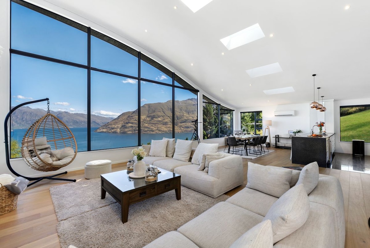 A bright living area features large windows that frame stunning views of the lake and mountains. Comfortable sofas invite relaxation, while a unique hanging chair adds a cozy touch. The space is complemented by soft lighting and a modern dining area in the background.