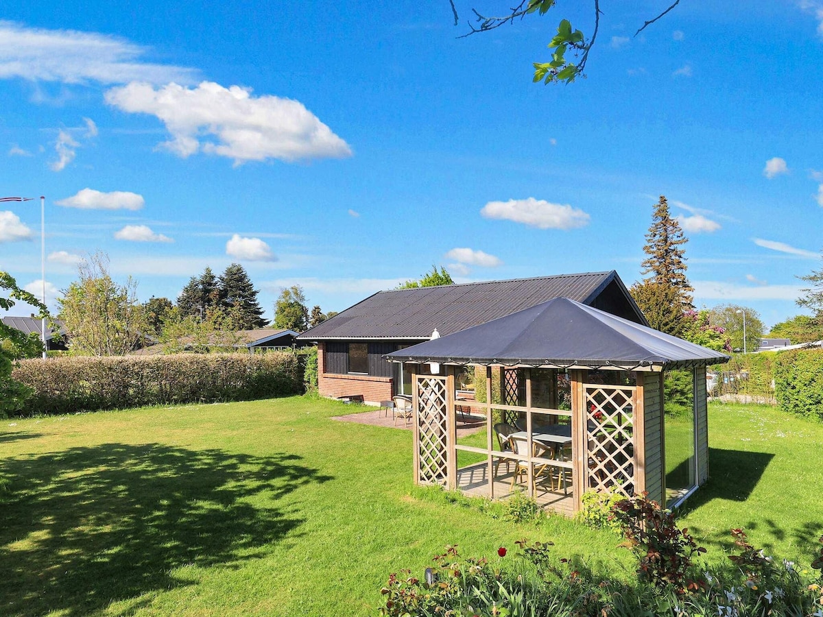 A spacious garden surrounds the holiday home, featuring a wooden gazebo with a slanted roof. Inside the gazebo, a dining area with tables and chairs is visible. Lush green grass and flowering plants create a vibrant outdoor space, with clear blue skies and a few clouds above.