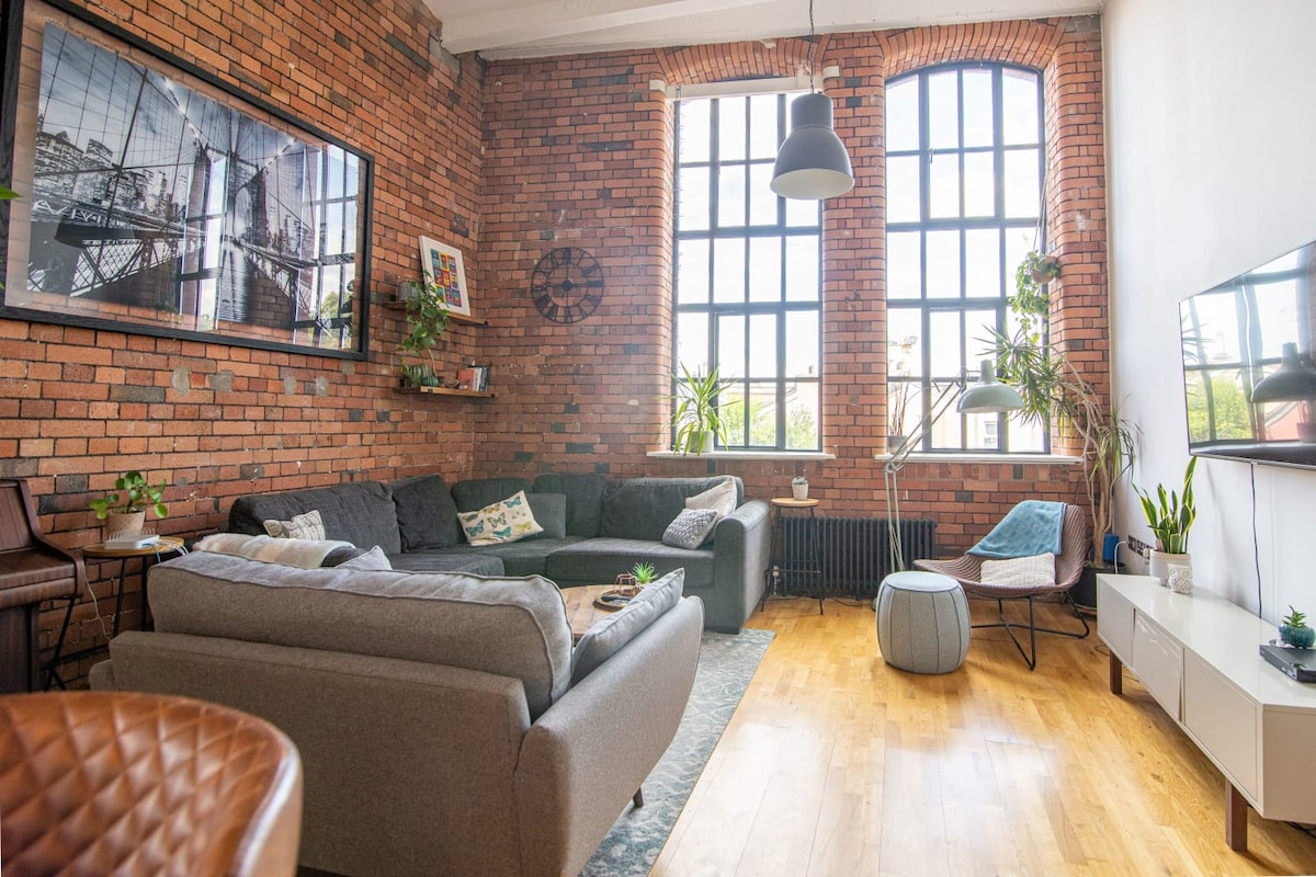 The spacious lounge features exposed brick walls and a double-height ceiling, allowing natural light to fill the room. Comfortable seating arrangements are highlighted, along with a large smart TV. Greenery is incorporated through various plants, creating a relaxed atmosphere.