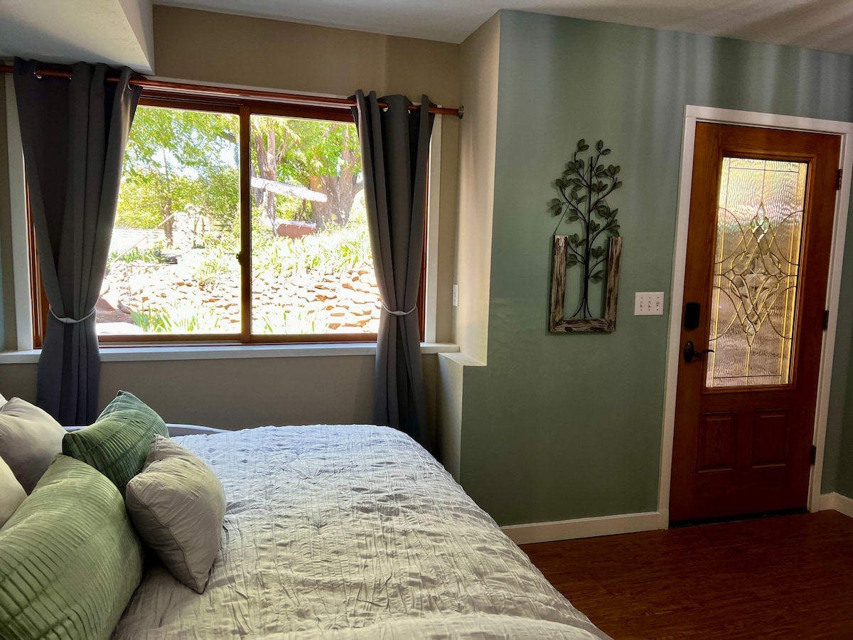 A well-lit corner of the room features a spacious bed adorned with soft pillows. A large window offers a view of the garden, framed by sheer gray curtains. A decorative wall art piece adds a touch of nature, while the entry door invites guests inside.