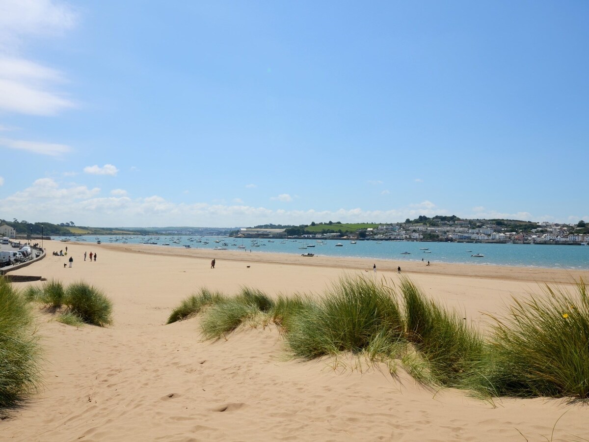 A sandy beach is visible, with gentle dunes of grass leading to the water's edge. Small boats are scattered across the calm blue sea. The distant village is characterized by white buildings, framed by green hills under a bright blue sky.