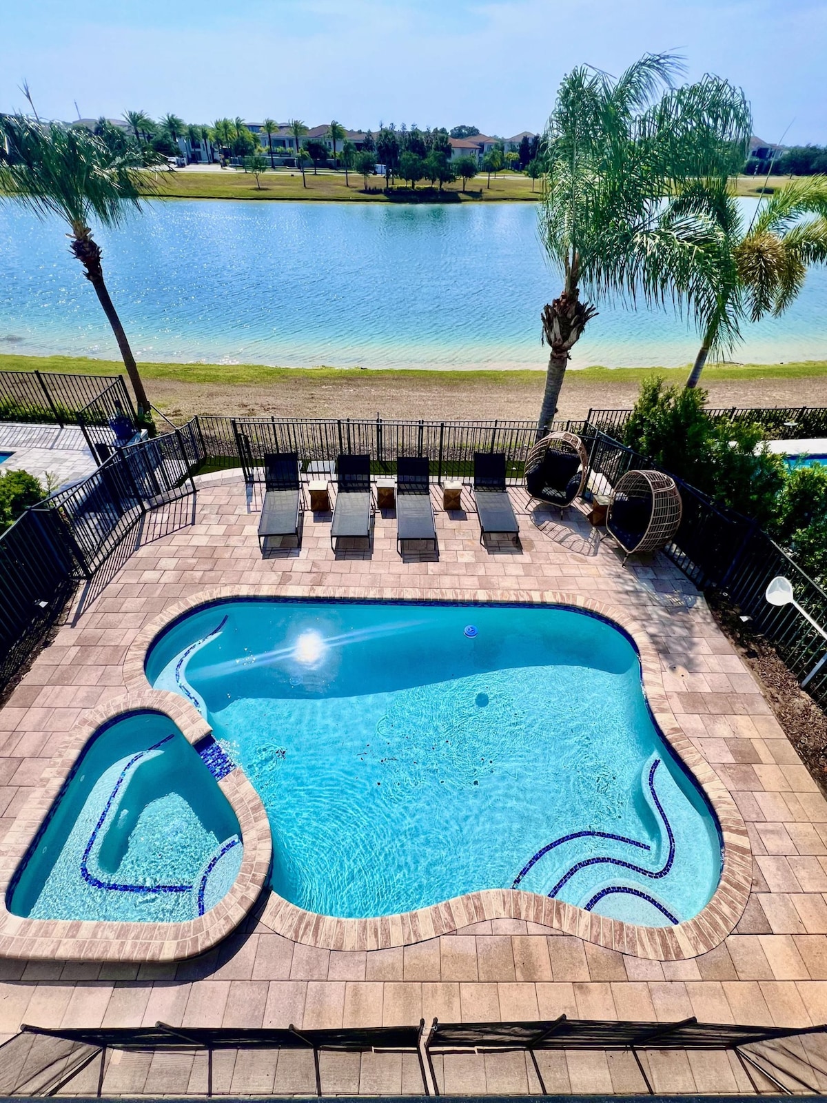 An outdoor space showcases a private pool surrounded by a spacious patio. Lounge chairs line the perimeter, and palm trees provide a tropical ambiance. The shimmering water reflects sunlight, while a serene lake is visible in the background, enhancing the tranquil atmosphere.