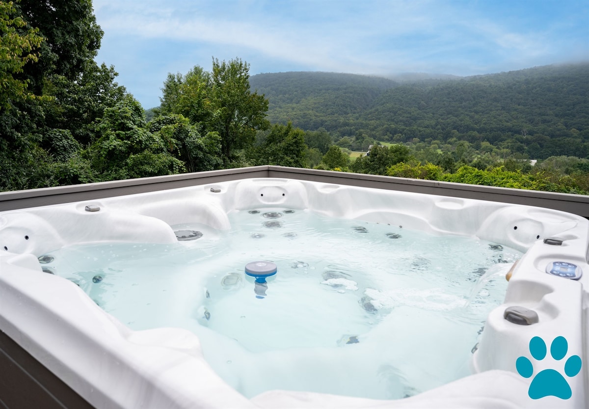 A hot tub is situated on a wooden deck, providing a relaxing space with scenic views of the surrounding lush green hills. Water glistens under natural light, creating an inviting atmosphere for a tranquil experience.