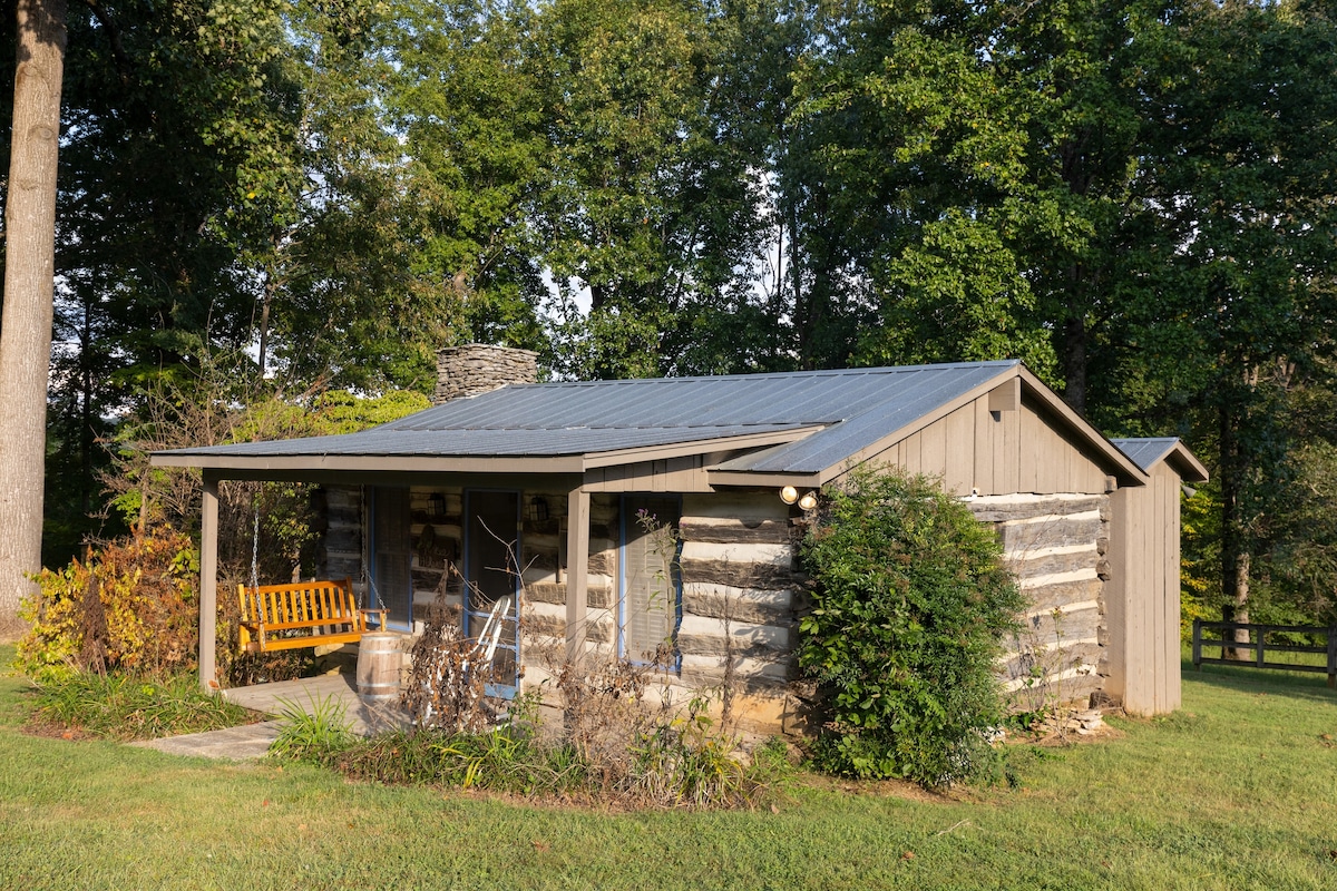 A cozy cabin featuring a rustic log exterior is set amid lush greenery. A covered porch is visible, adorned with a wooden swing and surrounded by colorful landscaping. The structure showcases a combination of natural logs and modern materials under a metal roof.