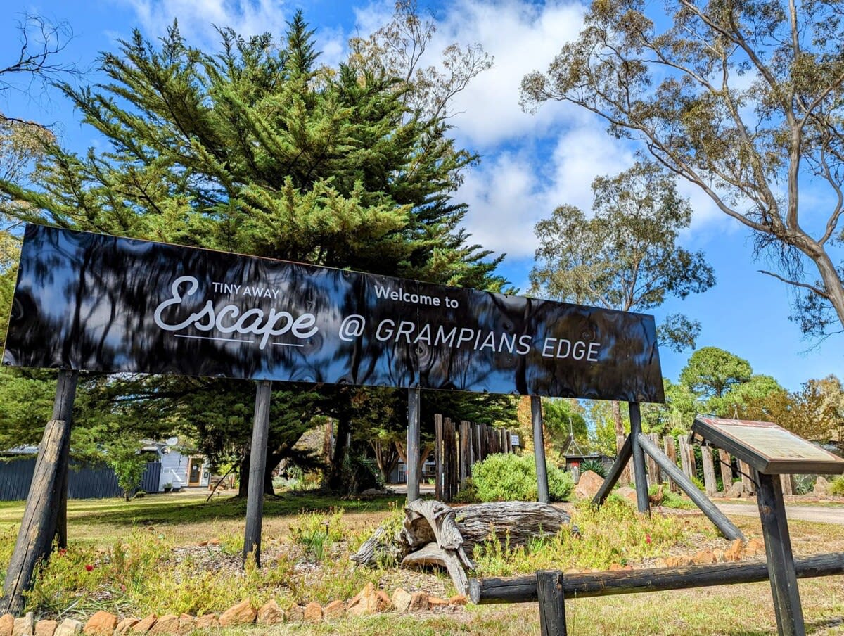 A welcoming sign for Tiny Away Escape at Grampians Edge is prominently displayed among lush trees. The clear sky is visible above, providing a serene backdrop. The sign features white text against a black background, indicating the location to guests arriving at the park.