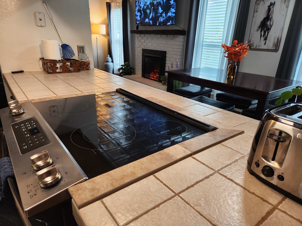 A spacious kitchen features a modern stovetop integrated into a tiled countertop. A digital display is visible above the stovetop, and a toaster stands nearby. A dining table can be seen in the background, along with a fireplace adorned with decorative elements and a television mounted on the wall.