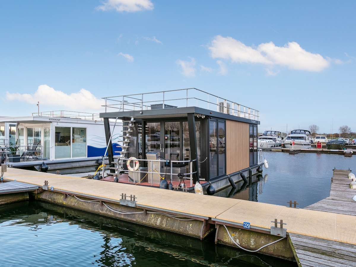 A modern houseboat is anchored at the harbor, showcasing large windows and a spacious roof terrace. The exterior features a combination of wood and glass elements. Nearby boats are visible, and the calm waters reflect the clear blue sky, creating a serene atmosphere.