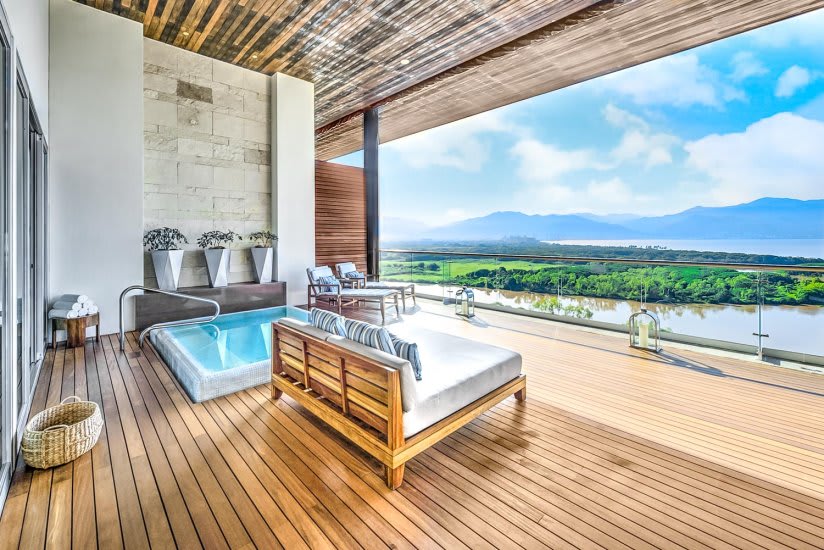 A spacious terrace features a heated splash pool and a king-size bed surrounded by plush lounge chairs. The wooden decking extends outward, offering expansive views of the lush landscape and distant mountains.