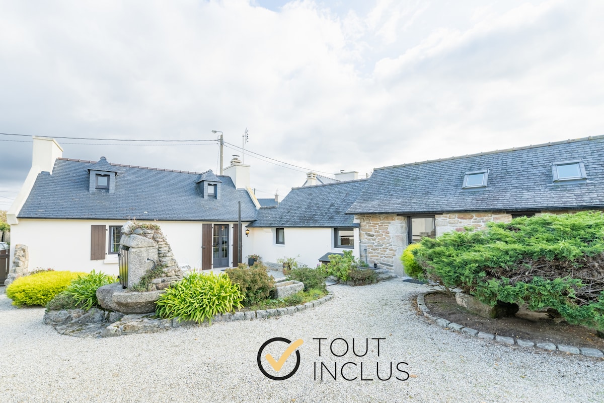 The exterior of a charming Breton house is visible, featuring stone walls and a slate roof. A lush garden surrounds the structure, with well-maintained greenery and a decorative stone well in the foreground. The property has a gravel pathway leading to the entrance.