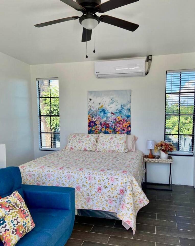 A bright sleeping area features a spacious bed covered with a floral-patterned blanket. Two windows allow natural light to fill the room, and an air conditioning unit is mounted on the wall. A small sitting area is equipped with a blue couch and a decorative pillow.