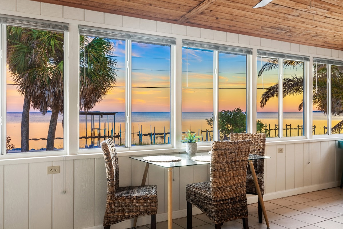 A dining area is positioned by large windows, providing expansive views of the Gulf at sunset. Two wicker chairs and a glass-topped table offer a comfortable space to enjoy meals while observing the vibrant colors of the sky and the serene water.