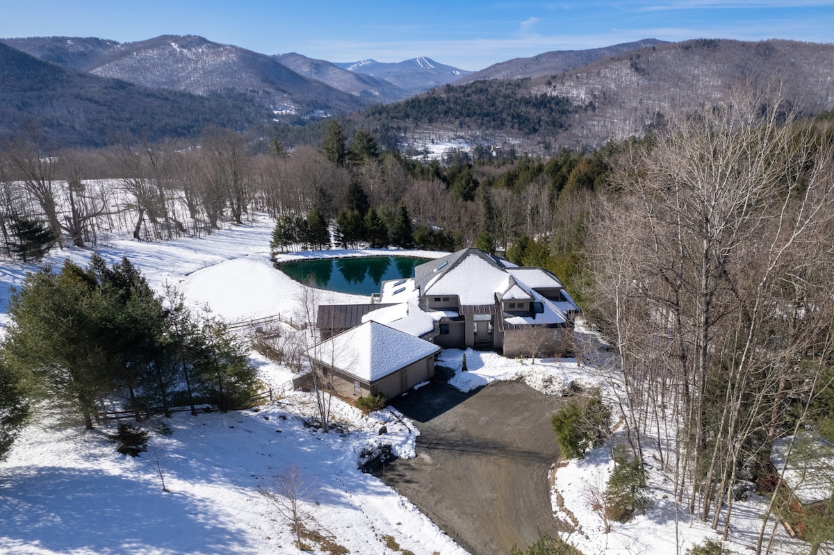 An aerial view showcases the expansive 10-acre estate surrounded by snow-covered mountains. The property features a serene, spring-fed pond reflecting the landscape, with a spacious home nestled near the water, offering an inviting retreat in a tranquil setting.