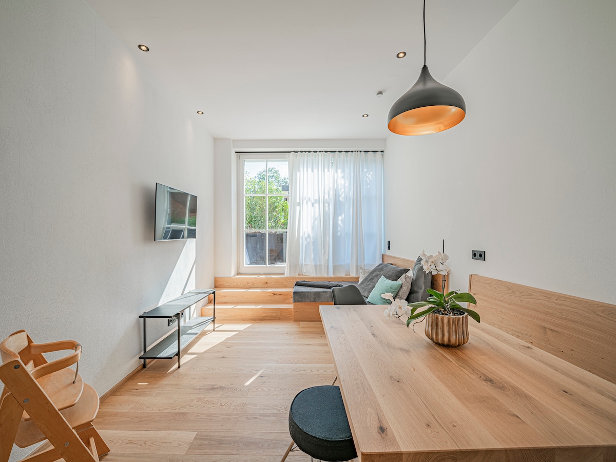 A bright living/dining area features an abundance of natural light from large windows. A comfortable seating area with plush cushions sits next to a wooden dining table, adorned with a small potted plant. A television is mounted on the wall, complementing the modern design.
