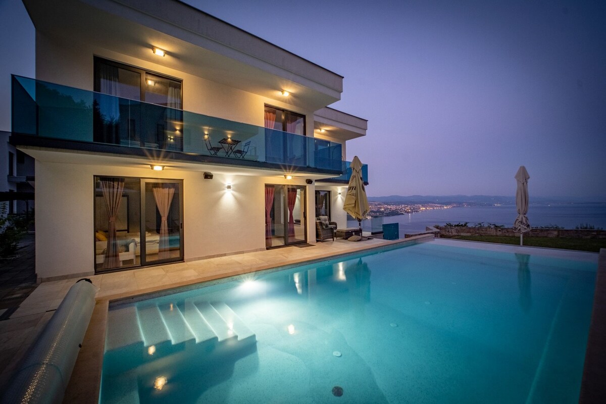 The modern villa is illuminated at dusk, showcasing a spacious outdoor pool and covered terrace. Large glass doors provide access to the interior, revealing hints of the inviting living spaces. The panoramic view of Kvarner Bay enhances the serene atmosphere.