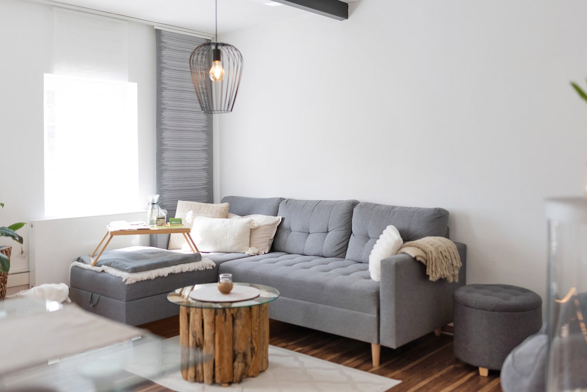 A modern living area features a grey sectional sofa paired with a round coffee table made from natural wood. Decorative cushions add comfort, while a soft rug and plant enhance the welcoming atmosphere. A stylish pendant light hangs above, casting a warm glow across the space.