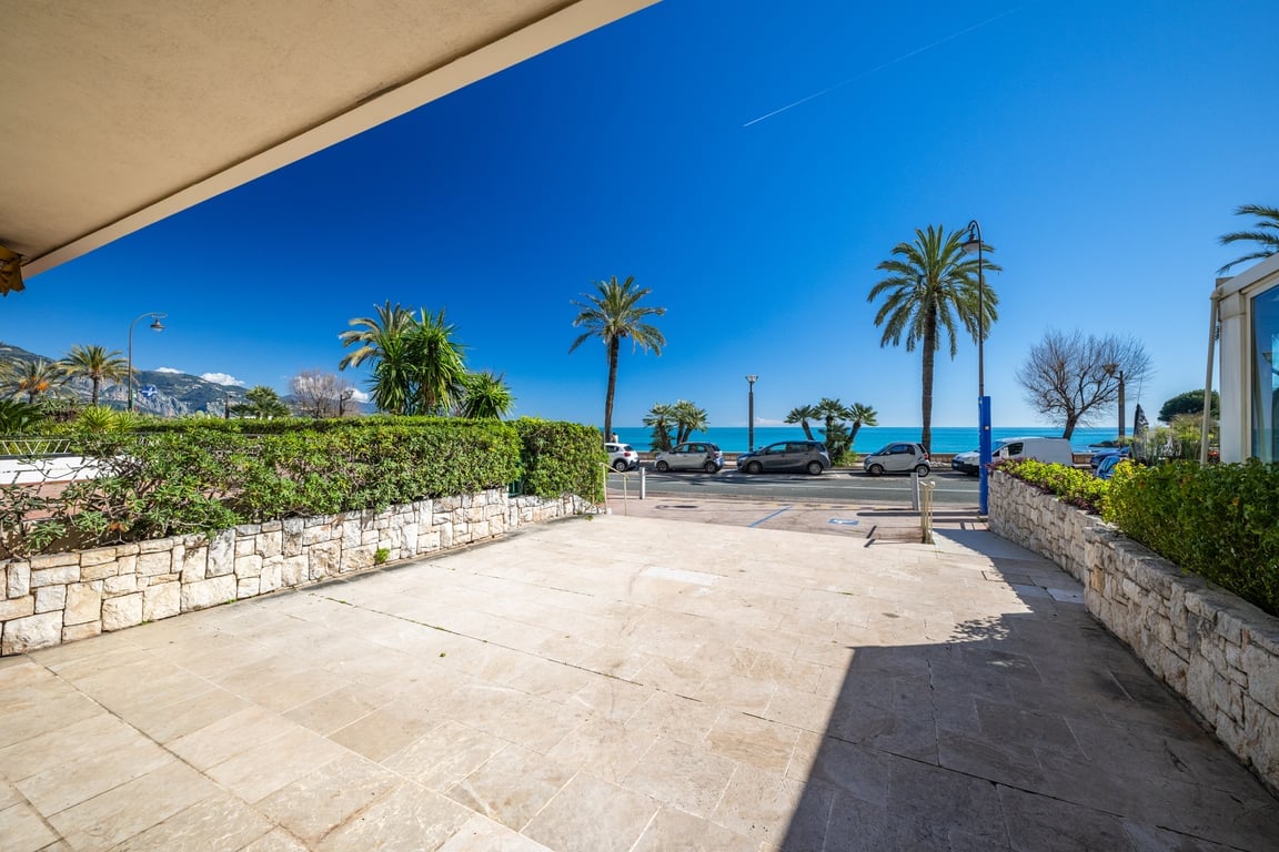 A spacious outdoor area features a stone pathway lined with greenery. Beyond the entrance, palm trees and the azure sea can be seen in the distance, creating a serene backdrop. The clear blue sky enhances the calming atmosphere.