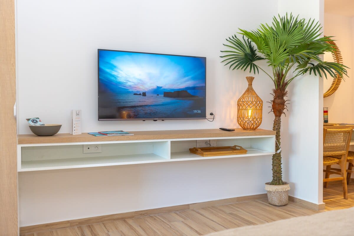 A modern entertainment unit hosts a flat-screen television displaying a serene beach scene. A decorative palm plant adds a natural touch, while a woven lantern provides soft lighting. The space features light wood accents, enhancing a clean, contemporary ambiance.