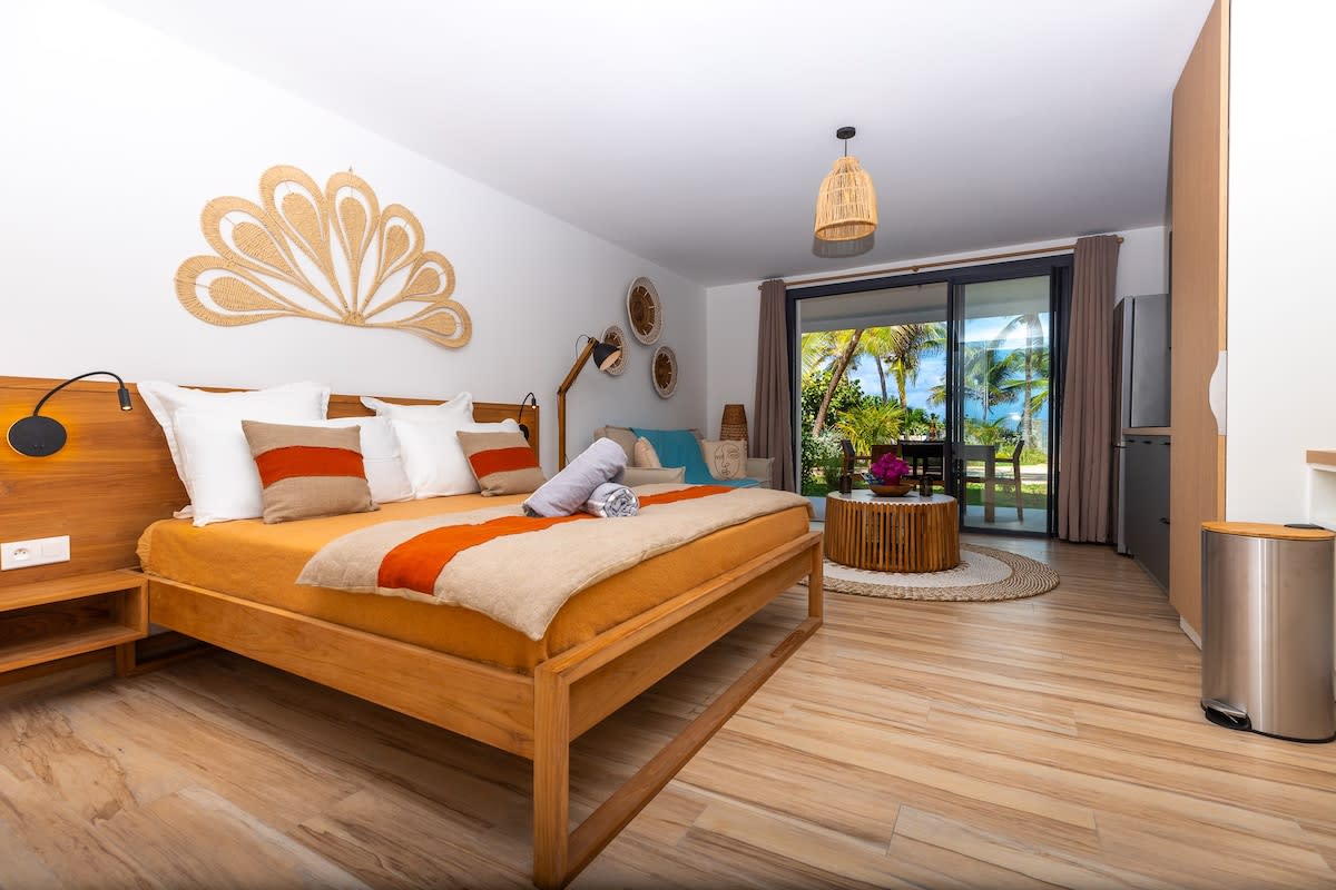 The studio features a spacious bed dressed in soft linens, complemented by decorative elements on the walls. Large glass doors offer views of a tropical landscape, while wooden accents and natural textures create a serene ambiance. A comfortable seating area is visible, enhancing the inviting space.