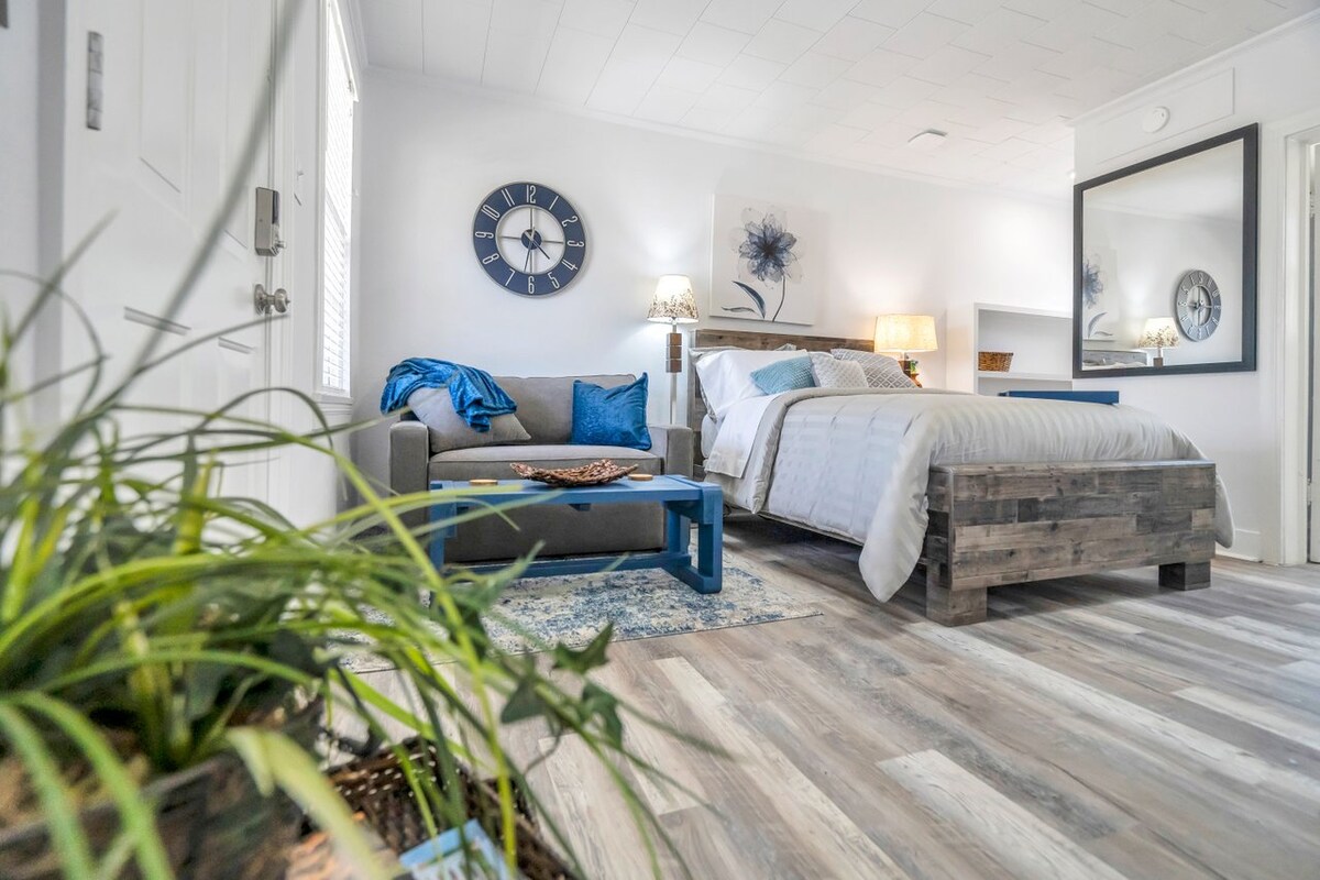 A well-arranged studio apartment features a cozy living area with a gray sofa and a blue accent table. A spacious bed is paired with neutral bedding and situated near a wall-mounted mirror. A plant adds a touch of greenery to the warm interior.