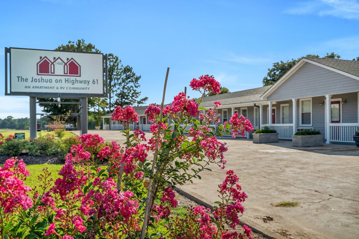 A welcoming view of the property exterior showcases a sign reading 'The Joshua on Highway 61.' Vibrant pink flowers in the foreground complement the well-kept area. The light-colored building features a covered porch area, contributing to the inviting atmosphere of the studio apartment.