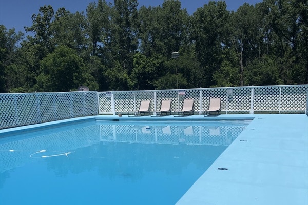 A serene private pool is displayed, featuring clear blue water that reflects the sunlight. Surrounding the pool, a white fence provides a boundary, while several lounge chairs are neatly arranged on the deck, inviting relaxation amidst a lush backdrop of greenery.